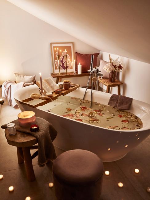 Biała wolnostojąca wanna wypełniona wodą i płatkami kwiatów, wokół drewniane dodatki jak stołek czy półka