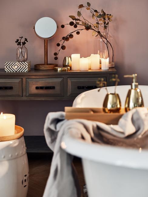 Drewniana konsola z szufladami w łazience na której stoi lustro i kosmetyki, na pierwszym planie biała wolnostojąca wanna ze złotymi akcesoriami łazienkowymi