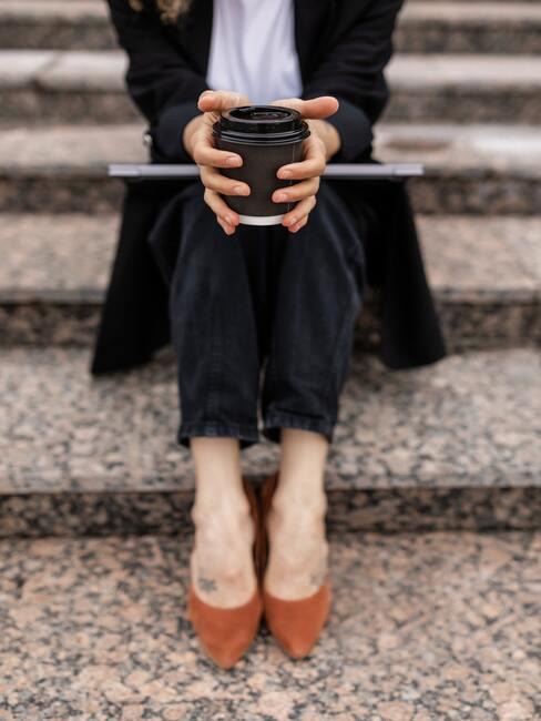 Kobieta w czarnym garniturze i kubkiem kawy siedząca na schodach