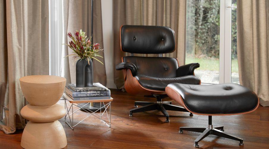 Eames lounge chair en voetenbankje naast een houten bijzettafel en ramen met beige gordijnen