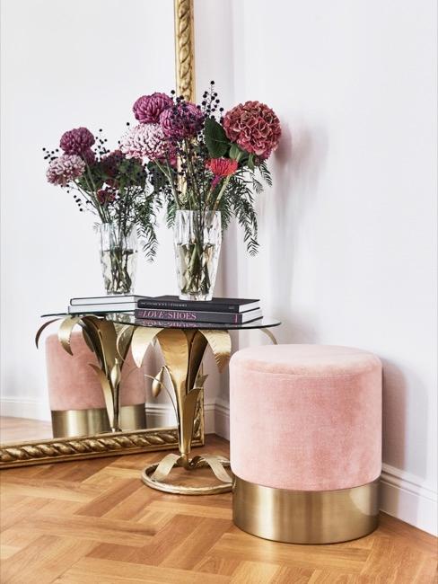 Tendances-banner Pouf rose et table basse en métal et verre avec deux livres sur lesquels se trouve un pot de fleurs, le tout placé devant un grand miroir