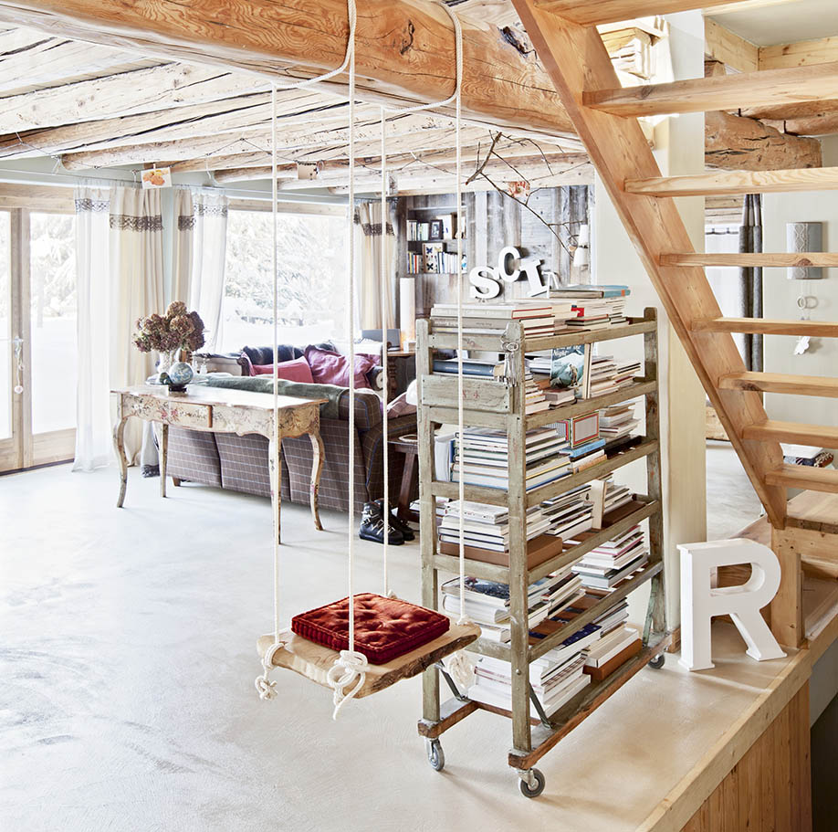 Child's swing hung from wooden beam next to bookshelves on castors in open-plan interior of wooden chalet